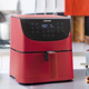 A cosori air fryer, red, sitting on a table amid several vegetables and pots and pans.